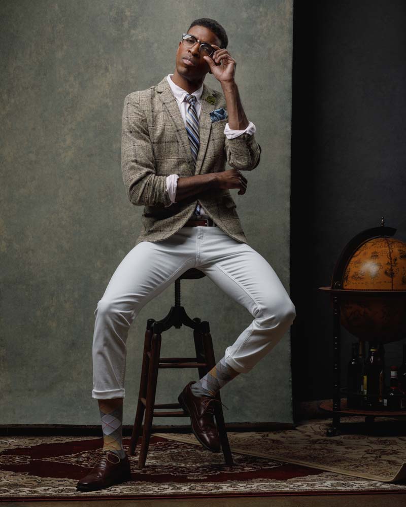 male model wearing a sophisticated outfit with glasses and tie sitting on chair