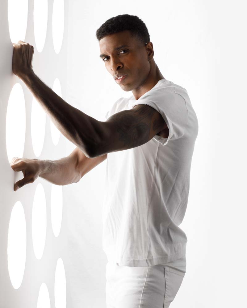 male model wearing all white standing next to wall with light shinning through holes