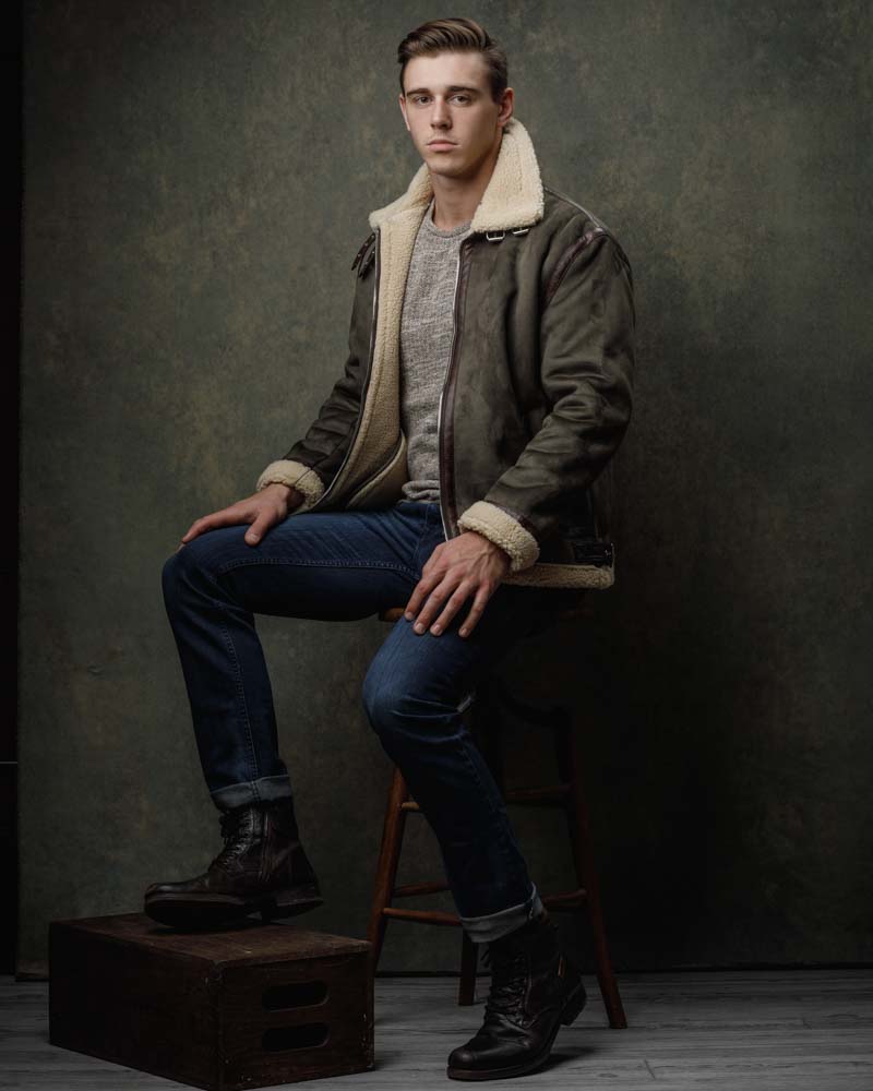 male model in front of dark green backdrop wearing dark green jacket with jeans and boots