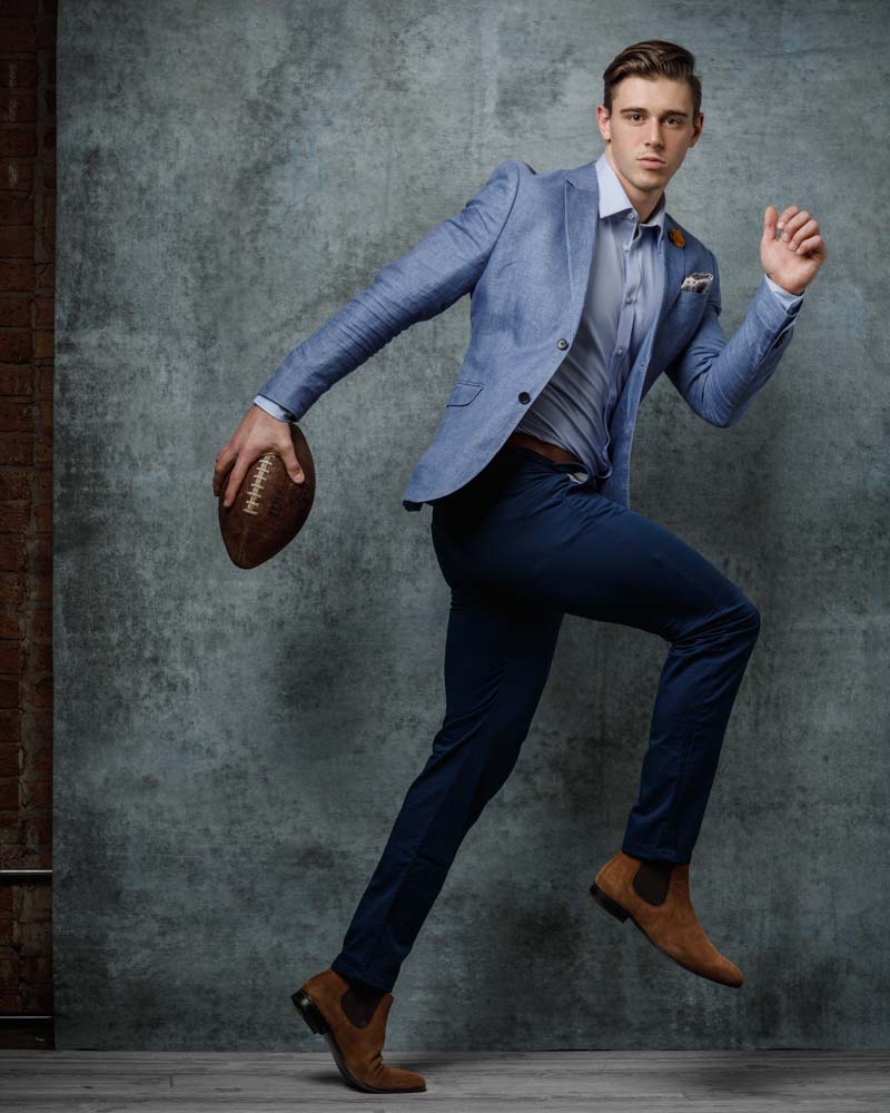  male model in front of rough light blue backdrop wearing an athletic suit and holding a football