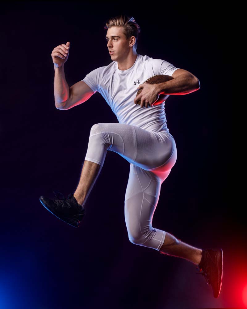  male model in front of rough light blue backdrop wearing an all white and holding a football