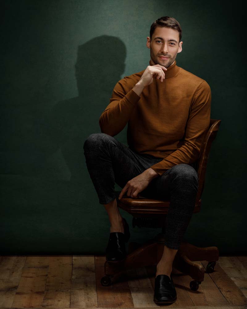 male model wearing a orange long sleeve turtle neck sitting on a chair in front of green background