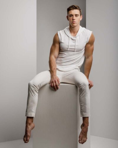 a muscular male model wearing an all white outfit/ the model is sitting on a white block with a geometrical background