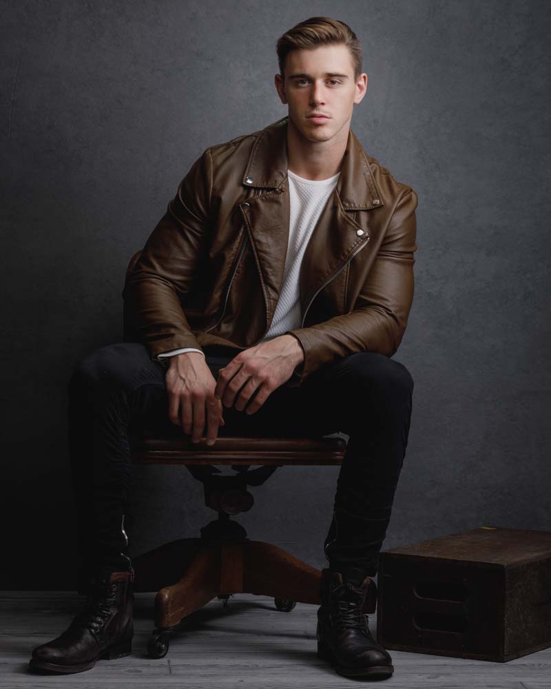 model is portrayed as a bad boy in a brown leather jacket and jeans in front of dark grey background