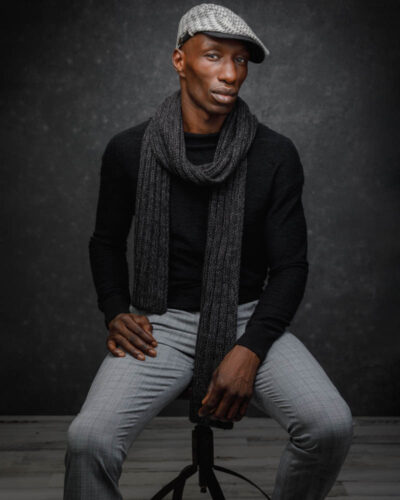 male model is dressed in fashionable black sweater and grey pants, the outfit is styled with a scarf and a hat, the model is posed confidently with the background textured dark grey