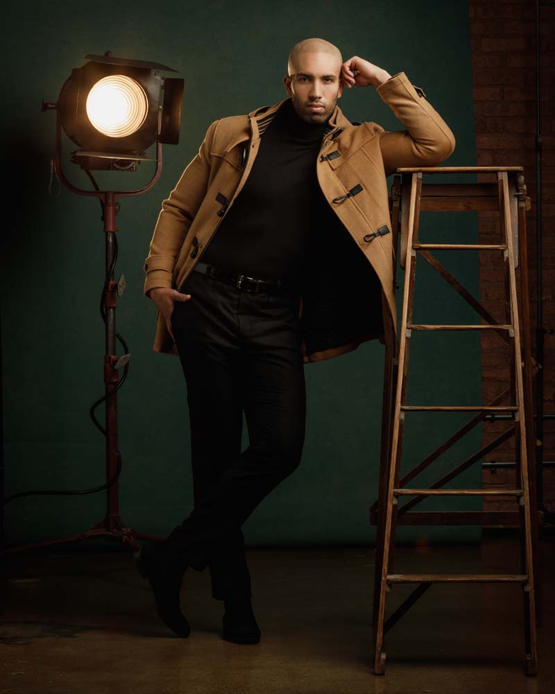 male model posed leaning on ladder wearing a tan jacket