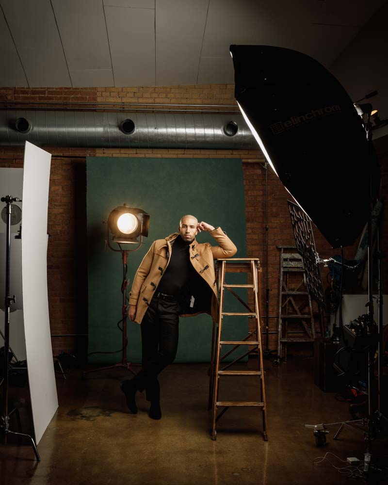 male model posed leaning on ladder wearing a tan jacket behind the scenes