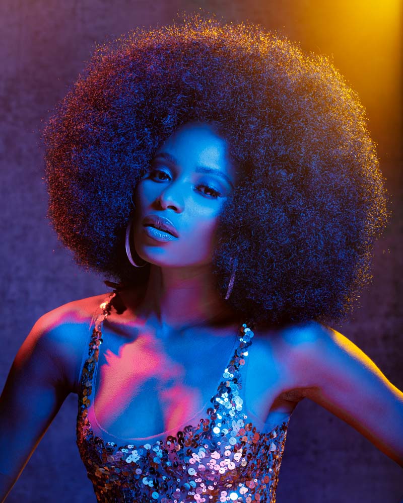  the models afro is styled in interesting shapes, female model with a round afro wearing a sparkly dress the model is illuminated with yellow, blue, and red light to create a disco party atmosphere