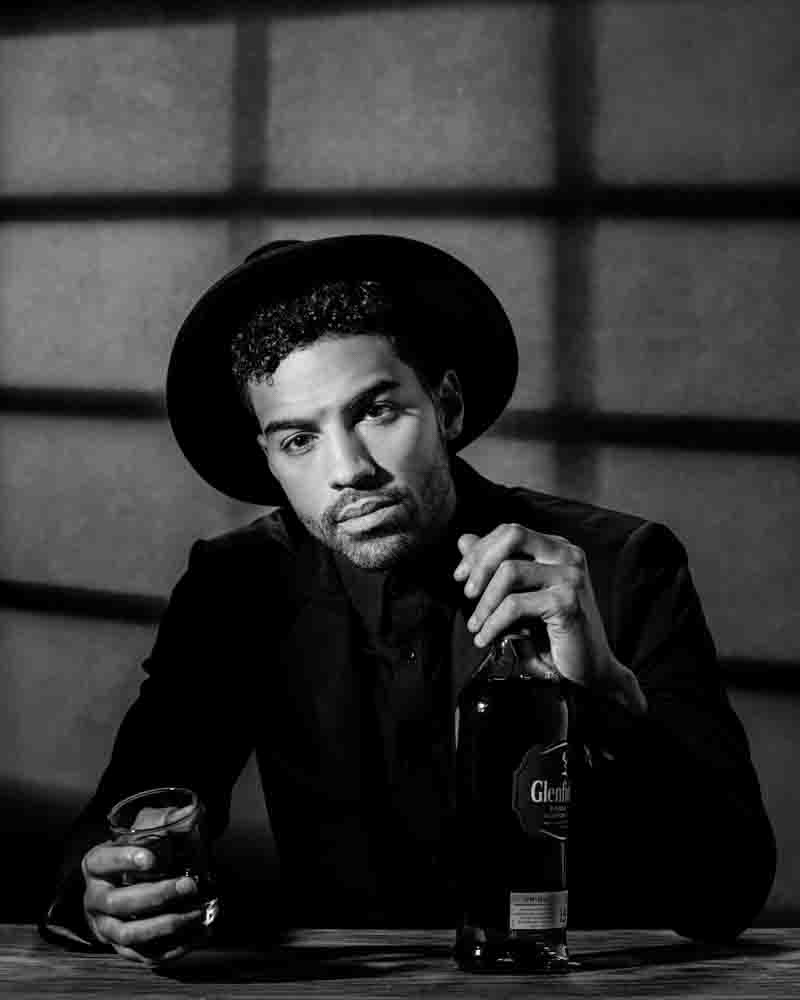 Cinematic black and white portrait of Giovanni holding a whiskey bottle, creating a moody, film-noir inspired look.The lighting in this portrait plays a critical role in crafting the mood. The shadows cast across Giovanni’s face and the background mimic the chiaroscuro technique often seen in classic cinema, particularly film noir. This style of lighting enhances the dramatic effect, making the portrait not only a photograph but a scene straight out of a movie.