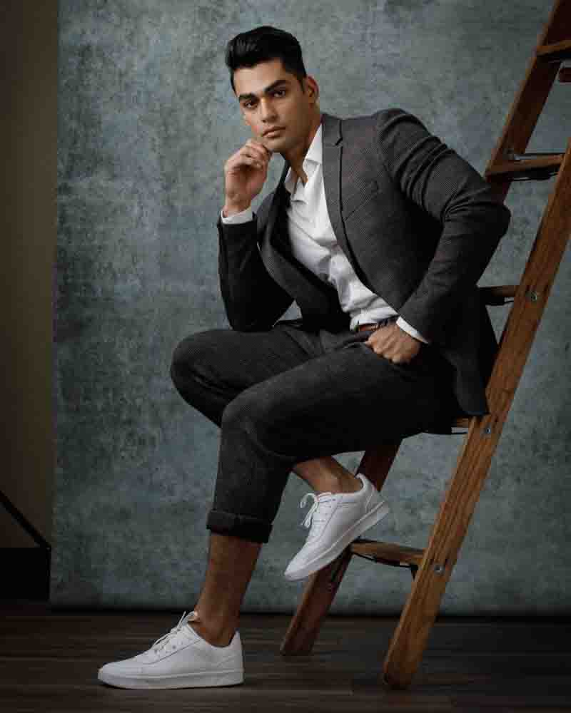 Akshay exudes a stylish mix of sophistication and laid-back charm, seated on a vintage wooden ladder against a muted blue-gray canvas, reflecting the eclectic vibe of Chicago's fashion landscape.