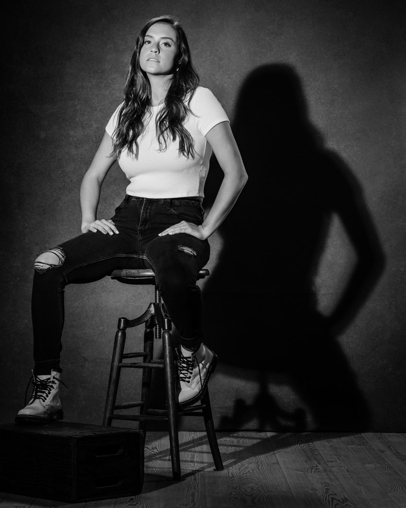 Taylor seated on a wooden stool, shadow cast on gray backdrop