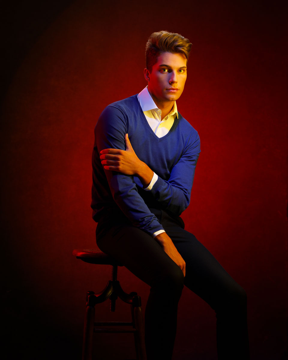 Three-quarter length portrait of Christian sitting in a blue sweater in front of a red background