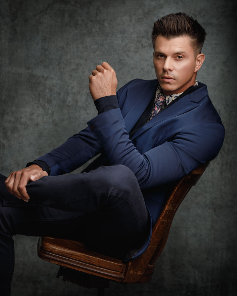 Kenning leaning back in a chair while wearing dark jeans and a blue suit jacket, 3/4 length chicago photographers headshot