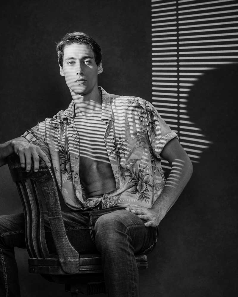 High contrast, black-and-white portrait of Marty in a partially opened floral print shirt, seated backwards on an antique wooden desk chair