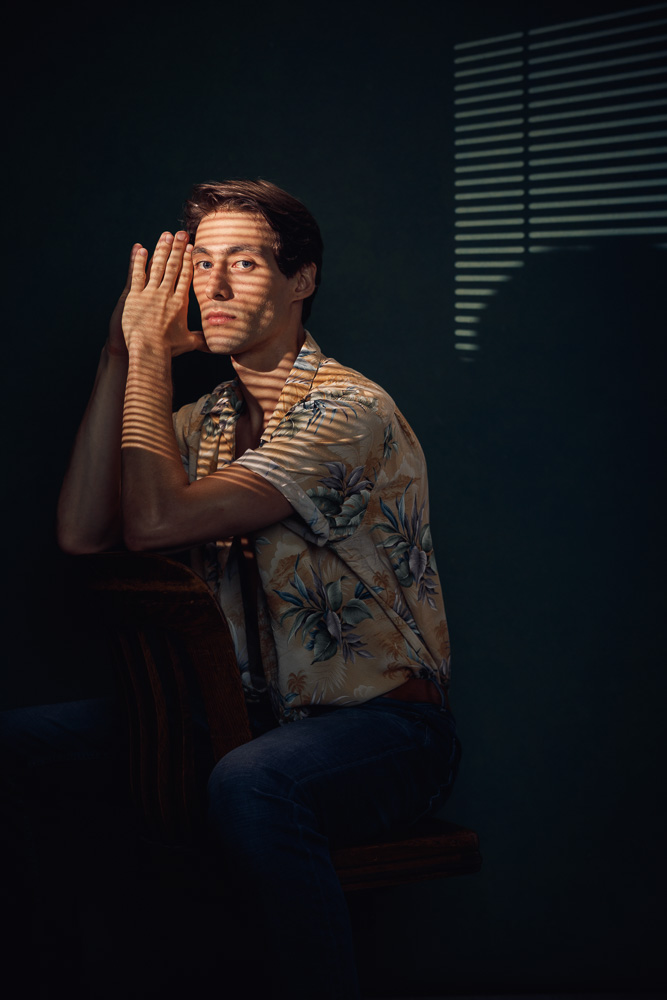 Marty in a partially opened floral print shirt, seated backwards on an antique wooden desk chair with light passing through mini blinds