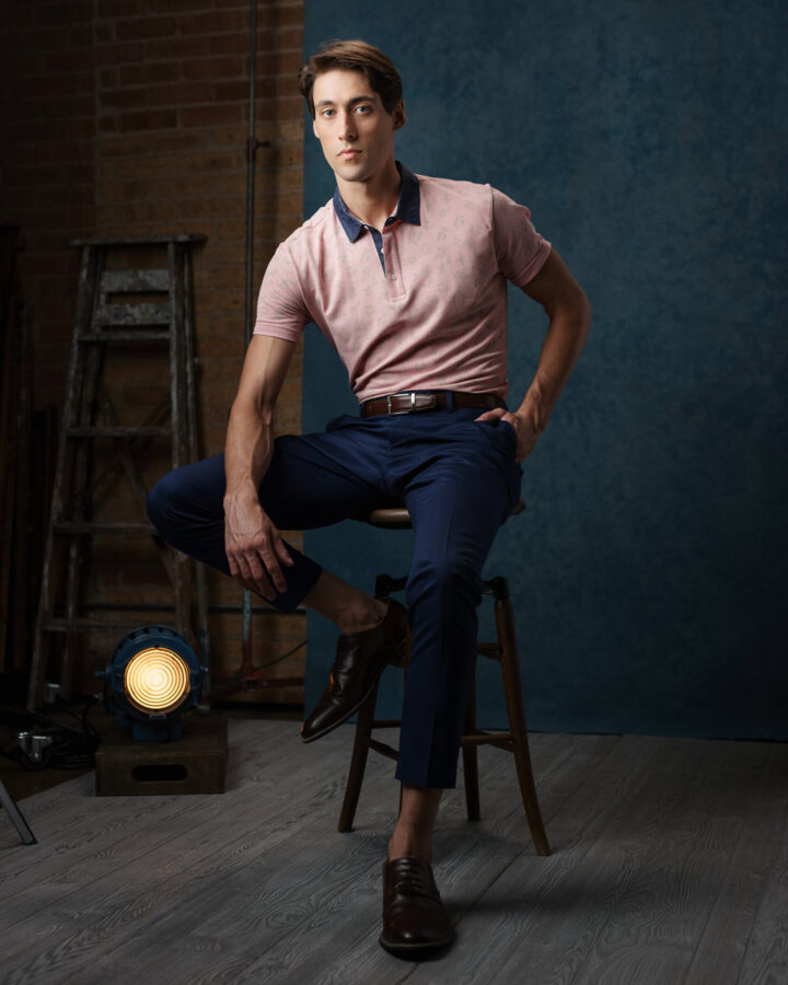 Editorial model photography in Chicago: Full body portrait of Marty, seated on an antique wooden stool with a vintage Hollywood movie light and ladder behind him. Wearing a blue and pink short sleeve polo, posing in front of a blue painted canvas backdrop.