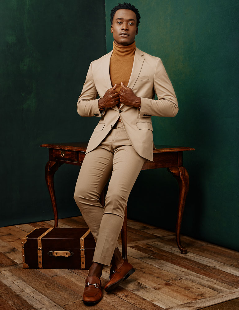 Model digital portfolio in Chicago - full length portrait of Justin in a tan suit with brown shoes and a brown turtleneck posing while seated on an antique wooden desk in front of two gray painted canvas backdrops.
