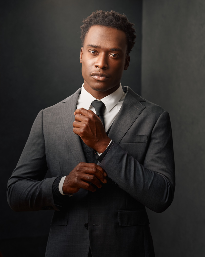 3/4 length portrait of Justin in a gray three-piece suit with a white shirt and a black tie in front of two gray canvas backdrops