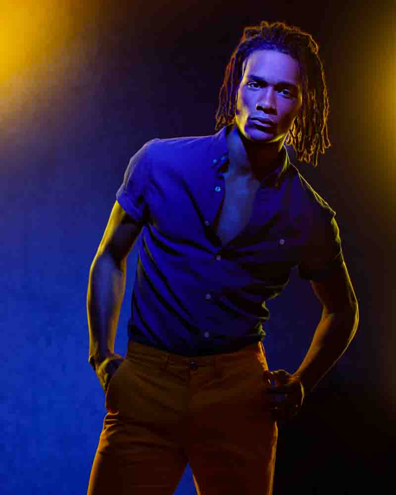 Matthew standing in a dramatic 3/4 length portrait wearing yellow pants and a partially open blue short sleeve button down shirt
