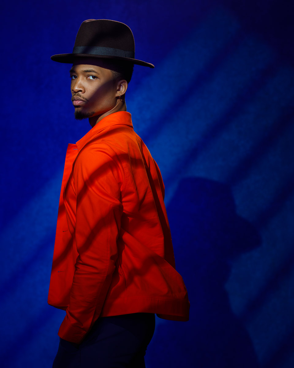Tyie in an orange jacket and brown hat, posing in front of a blue backdrop with creative lighting.