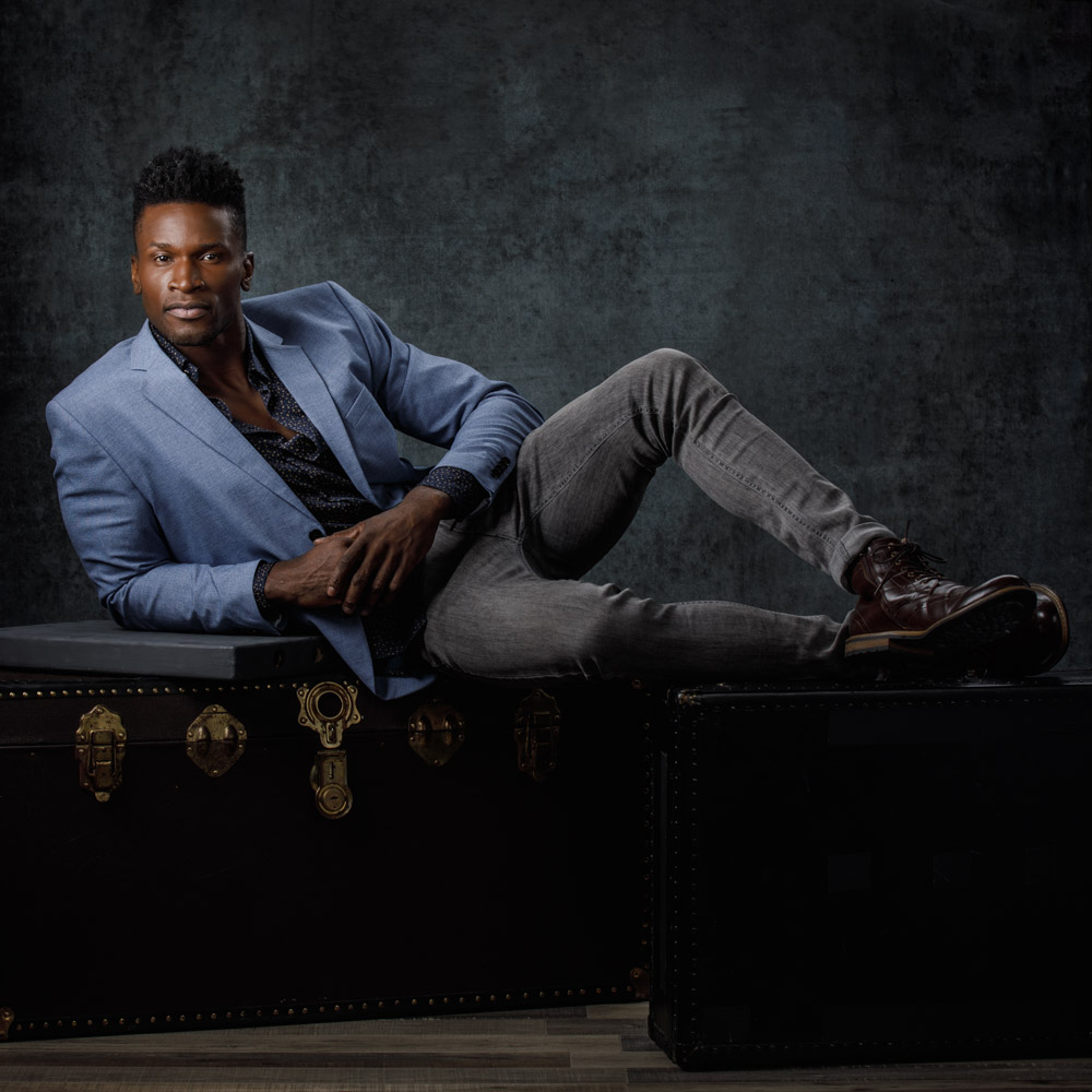 Chaun laying across antique luggage and trunk, dressed in stylish attire for his modeling portfolio service in Chicago