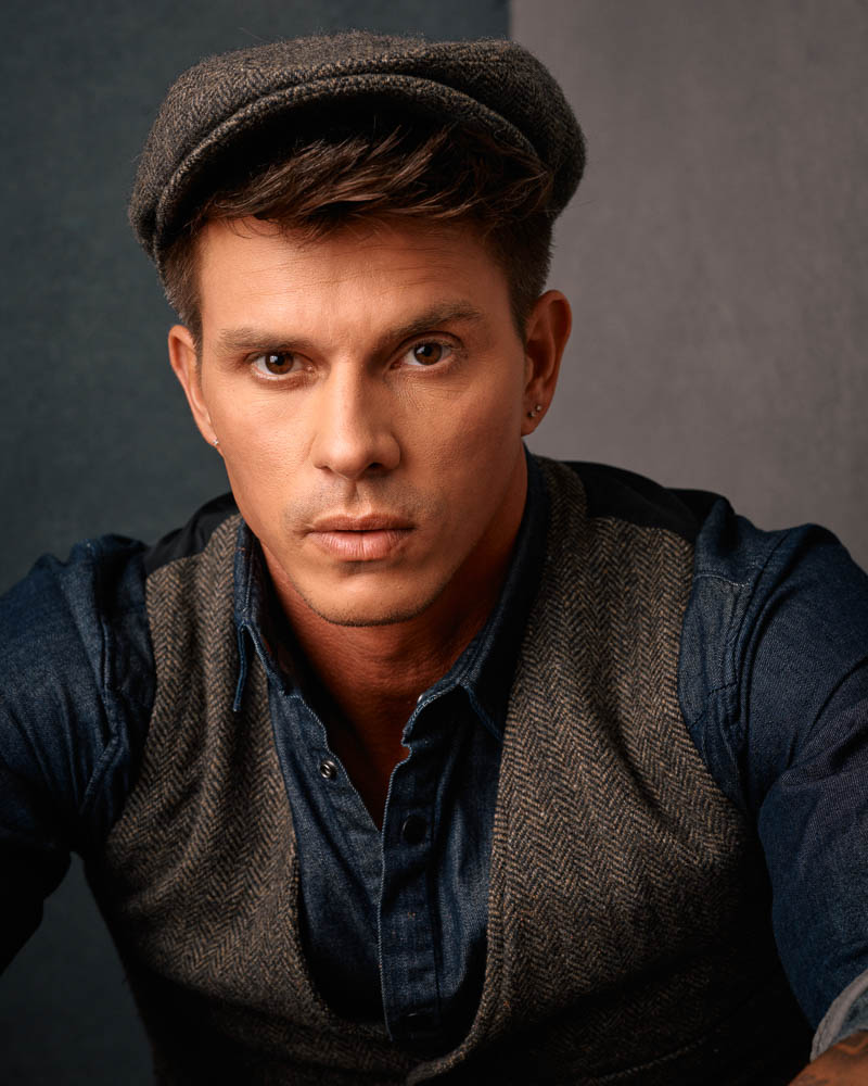This is a headshot of Kenny, sitting on an antique wooden stool in front of two painted canvas backdrops wearing a newsboy outfit complete with green pants, a blue denim shirt, a vest, and a cap