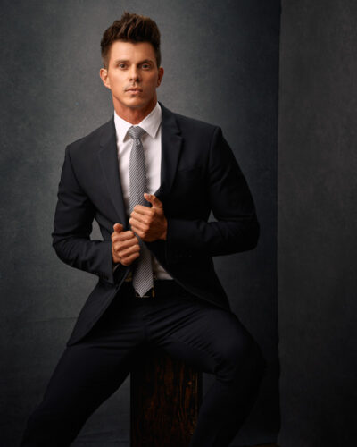 This is a waist up portrait of Kenny wearing a black suit with a white shirt and a gray tie in front of a gray painted canvas backdrop is an example of that you can expect from professional model photoshoot services in Chicago