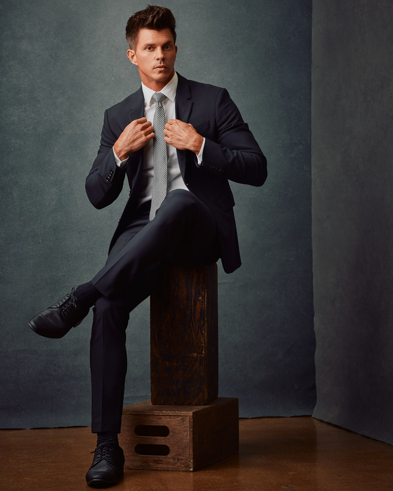 Kenny, wearing a black suit and tie, poses confidently against a gray painted canvas backdrop. This image exemplifies the sophisticated style captured in professional model photoshoots by John Gress Photography in Chicago.