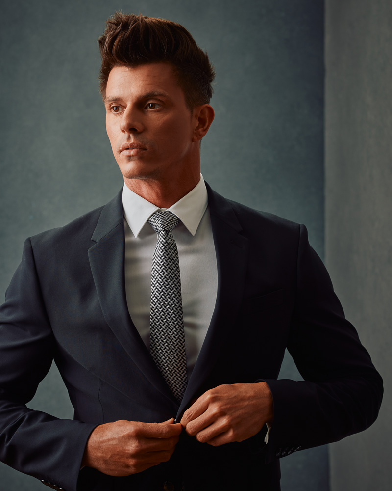 Kenny, in a black suit and tie, poses confidently against a gray painted canvas backdrop. This image exemplifies the sophisticated style captured in professional model photoshoots by John Gress Photography in Chicago.