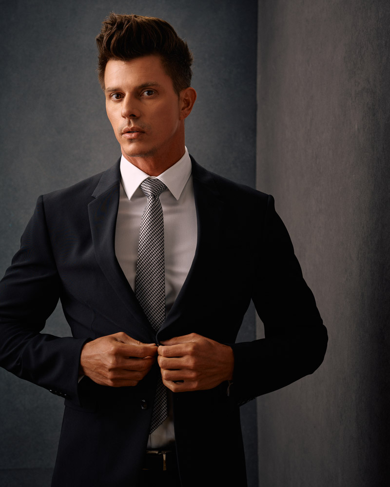 Kenny, wearing a black suit and tie, poses confidently against a gray painted canvas backdrop. This image showcases the professionalism of John Gress Photography in Chicago.