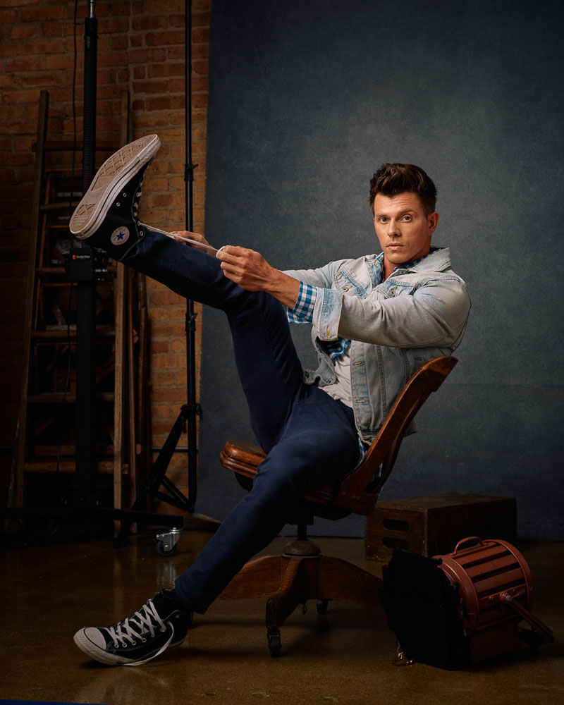 Kenny, in casual clothing, ties his shoes while leaning back in a wooden chair. This behind-the-scenes shot reflects the relaxed and professional atmosphere of a model photoshoot with John Gress Photography in Chicago.