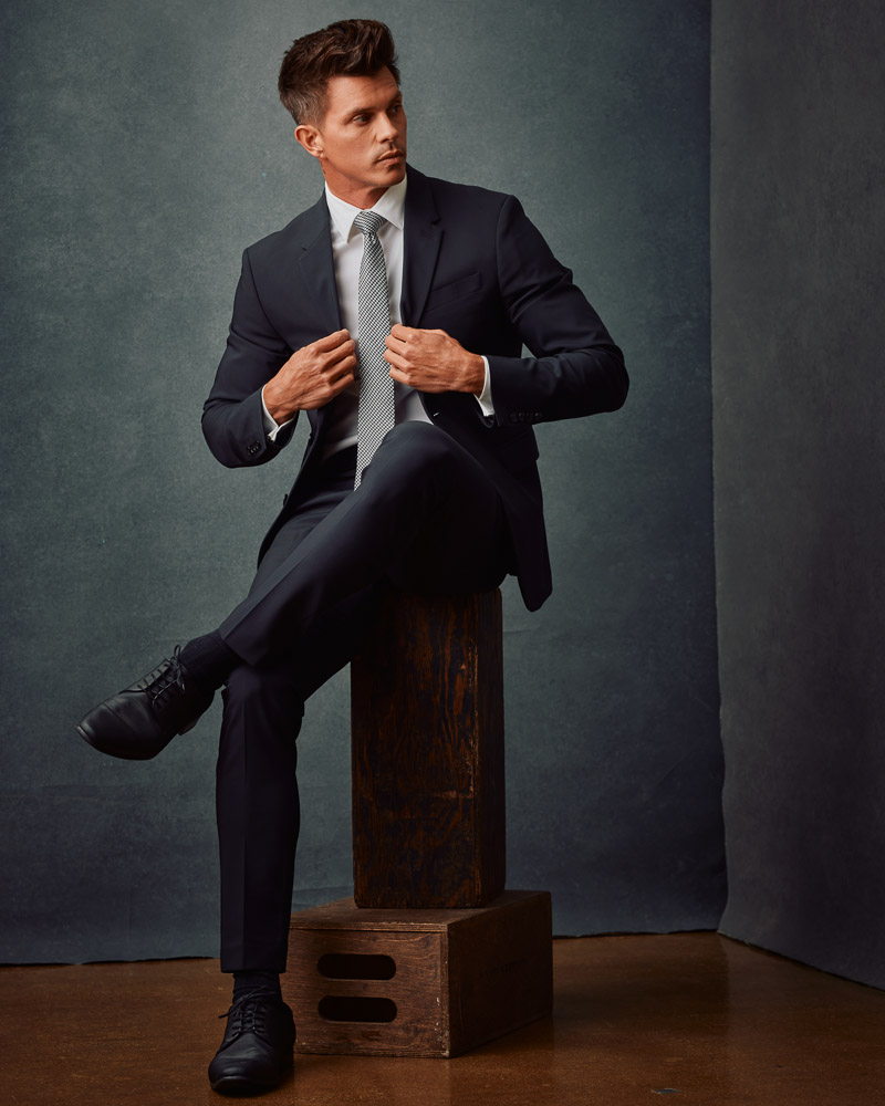 Kenny, wearing a black suit and tie, poses confidently against a gray painted canvas backdrop. This image exemplifies the sophisticated style captured in professional model photoshoots by John Gress Photography in Chicago.