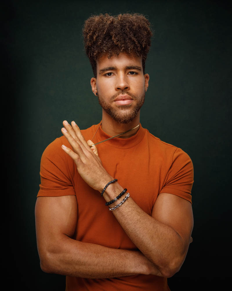 Trae, dressed in an orange shirt, poses in front of a green backdrop
