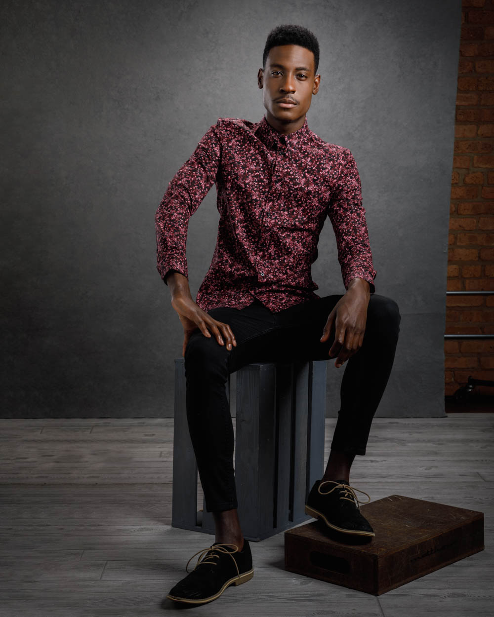 Full body portrait of David in black jeans and a maroon floral top, poised on a gray wooden crate