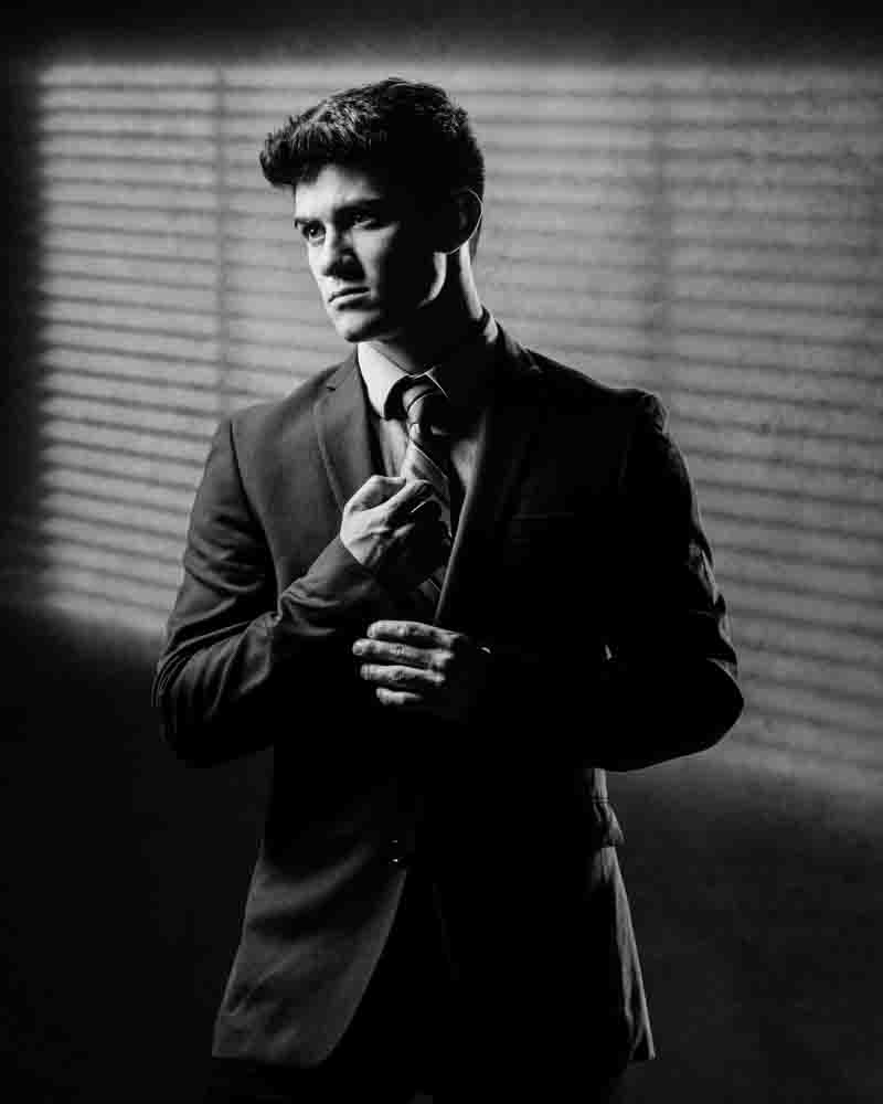 High contrast black and white character portrait of Kainoa in a suit, with shadow lines from miniblinds adding dramatic effect