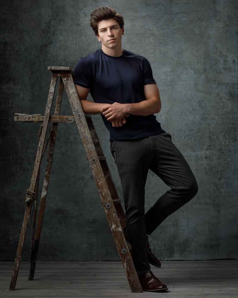 Full body portrait of Noah in gray dress pants and a fitted blue t-shirt, leaning on an antique ladder.
