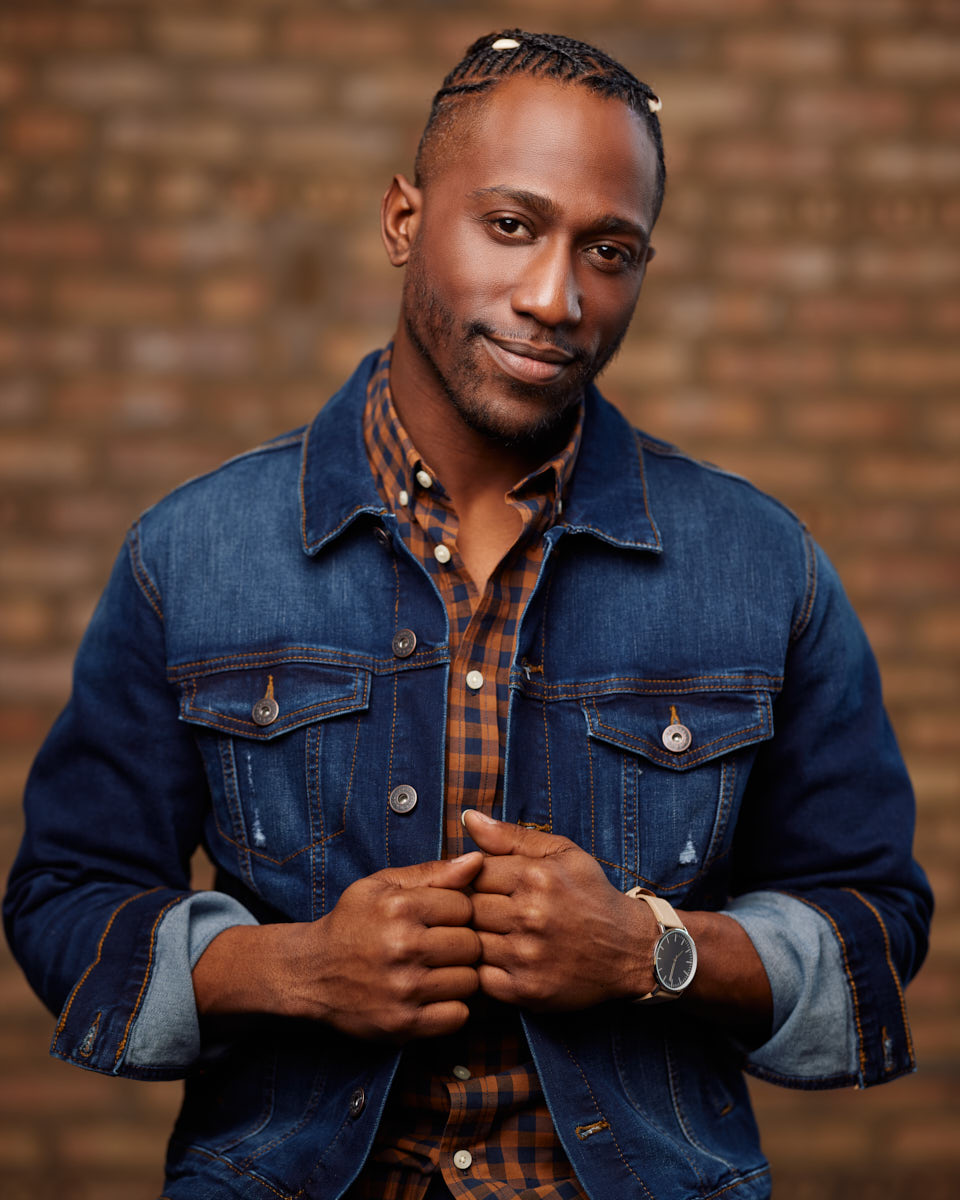 Preston is posing with a friendly smile, wearing a denim jacket over a checkered shirt. The background is a blurred brick wall, giving a warm tone to the image.