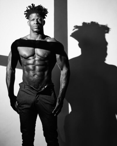 This is a three-quarter length shirtless portrait of Wes shot vertical in black-and-white with very high contrast lighting as sunlight stream, throw window, casting a shadow of the window frame by Chicago fitness model photographer