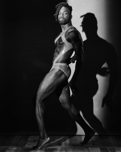 chicago fitness photography black and white image shows Amir standing full body in profile wearing a leatherette harness that also includes briefs. He is standing in a shaft of high contrast light and his shadow can be seen in the background on a painted canvass backdrop. The image really shows off his muscular layer, legs, abdominal muscle and arms