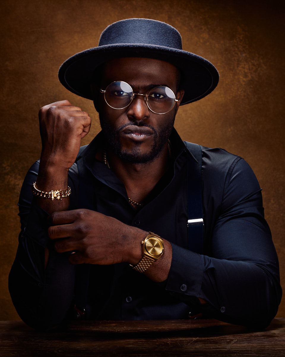 Dyren is wearing a black shirt and suspenders, accessorized with a gold watch and bracelet. He is also sporting a black hat and round glasses. He strikes a serious pose with his fist raised, highlighting the gold bracelet on his wrist.