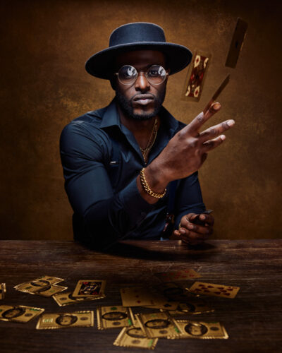 Dyren in a Chicago model photo studio. He is engaging in a dynamic action shot, flipping gold-colored playing cards into the air. The cards and more gold-colored money are scattered on the table. He keeps a focused expression, and his attire remains consistent with the previous images.