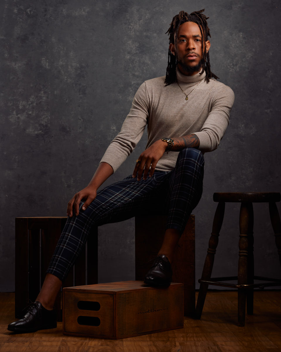 Jalen sits on wooden crates, wearing a light gray turtleneck sweater and plaid pants, exuding a sophisticated and stylish look.
