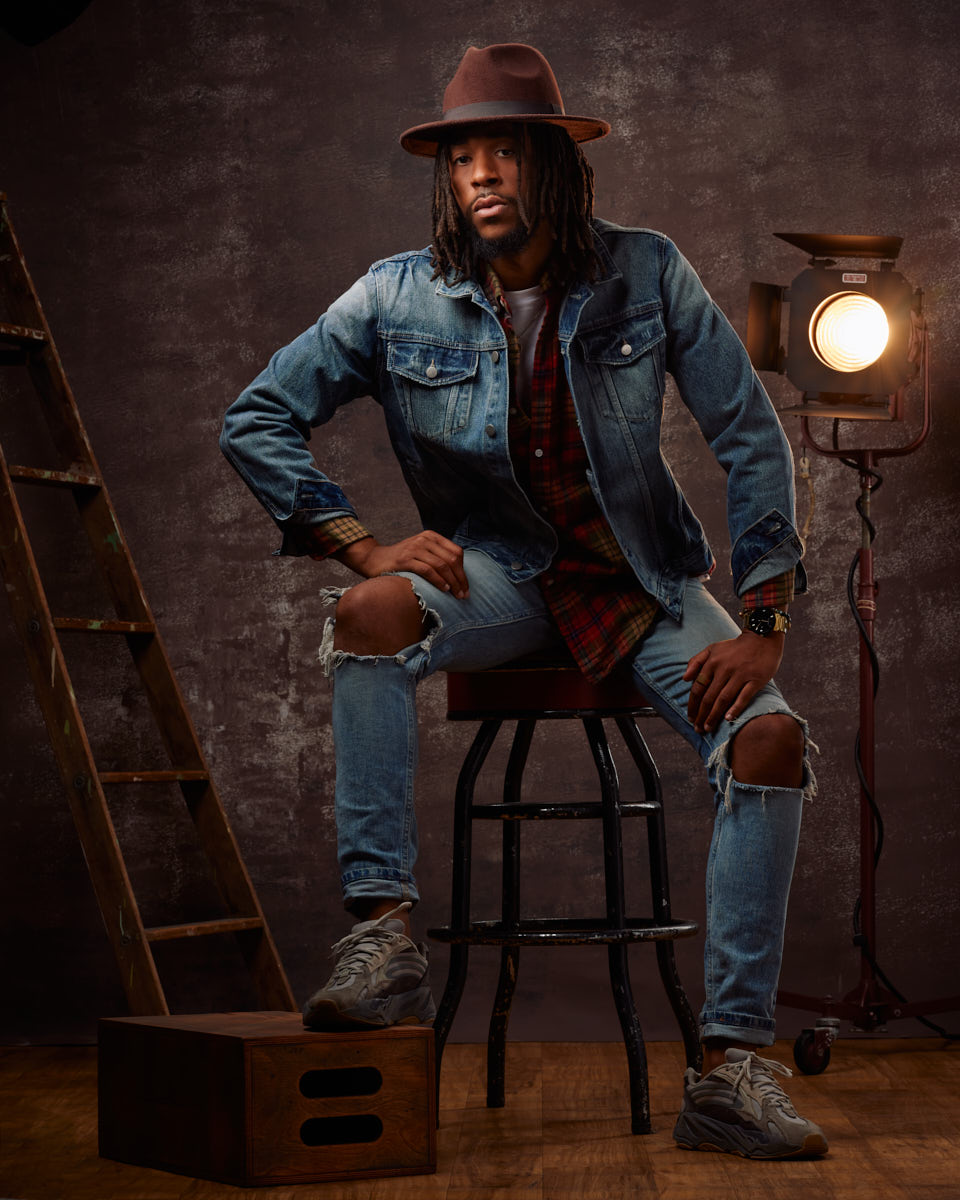 Jalen sits confidently on a stool in a Chicago model photo studio, wearing a denim jacket, distressed jeans, and a brown hat, with a rustic ladder and studio light in the background.