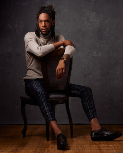 Jalen poses with his arms on the back of a chair in a Chicago model photo studio, wearing the same light gray turtleneck and plaid pants, creating a relaxed yet composed image.