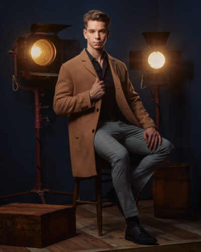 Erik seated in a long tan overcoat in front of vintage Hollywood lights and blue backdrops, exuding a timeless, cinematic feel by a Chicago modeling portfolio photographer