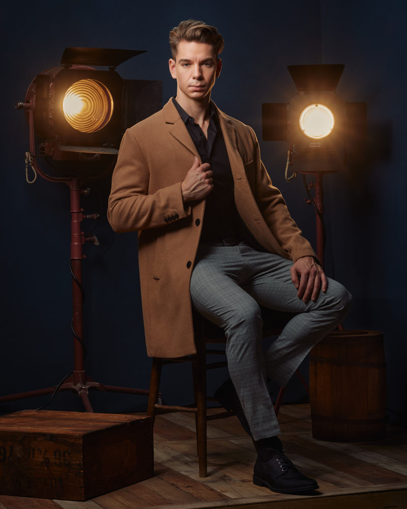 Erik seated in a long tan overcoat in front of vintage Hollywood lights and blue backdrops, exuding a timeless, cinematic feel by a Chicago modeling portfolio photographer