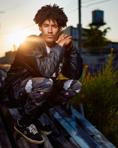 Chicago on-location male model photographer - Kasan crouches on the wooden planks, with his hands clasped, wearing a black jacket and camo pants, with the setting sun illuminating his face.
