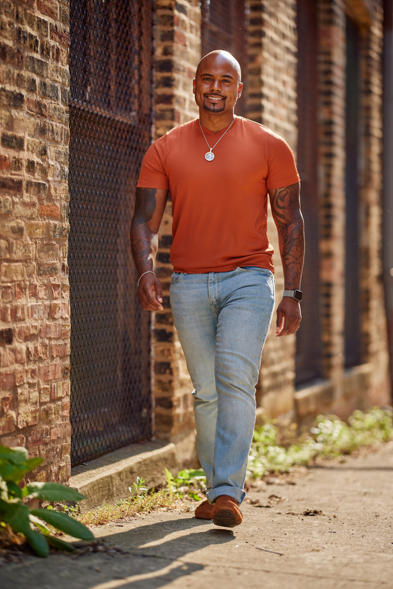 The model is captured walking along the same brick wall in the burnt orange t-shirt and light blue jeans, smiling as he approaches the camera.