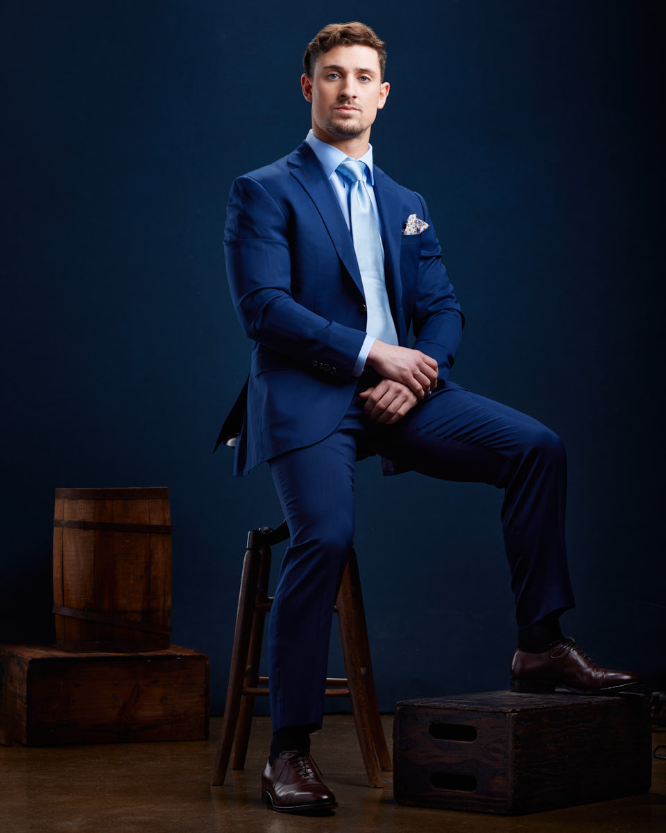 This image depicts a fitness model seated and dressed in a sharp blue suit with a light blue tie and pocket square. He is looking directly at the camera with a serious expression by a Fitness Model Headshots photographer in Chicago