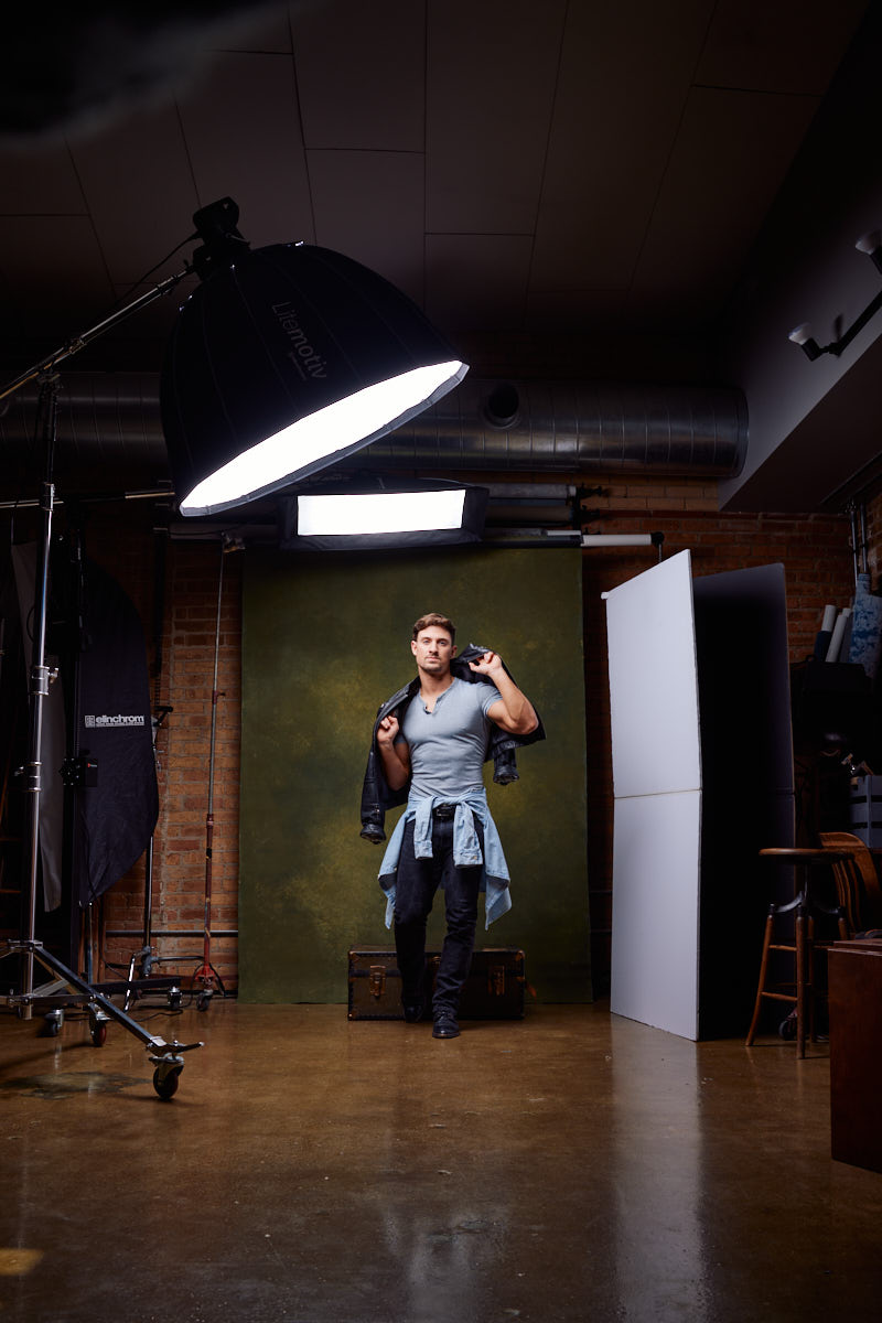 In this image, the fitness model is standing in a professional photography studio setup with lights and a backdrop. He is wearing a gray t-shirt, black jeans, and a denim jacket tied around his waist, holding a black jacket over his shoulder.